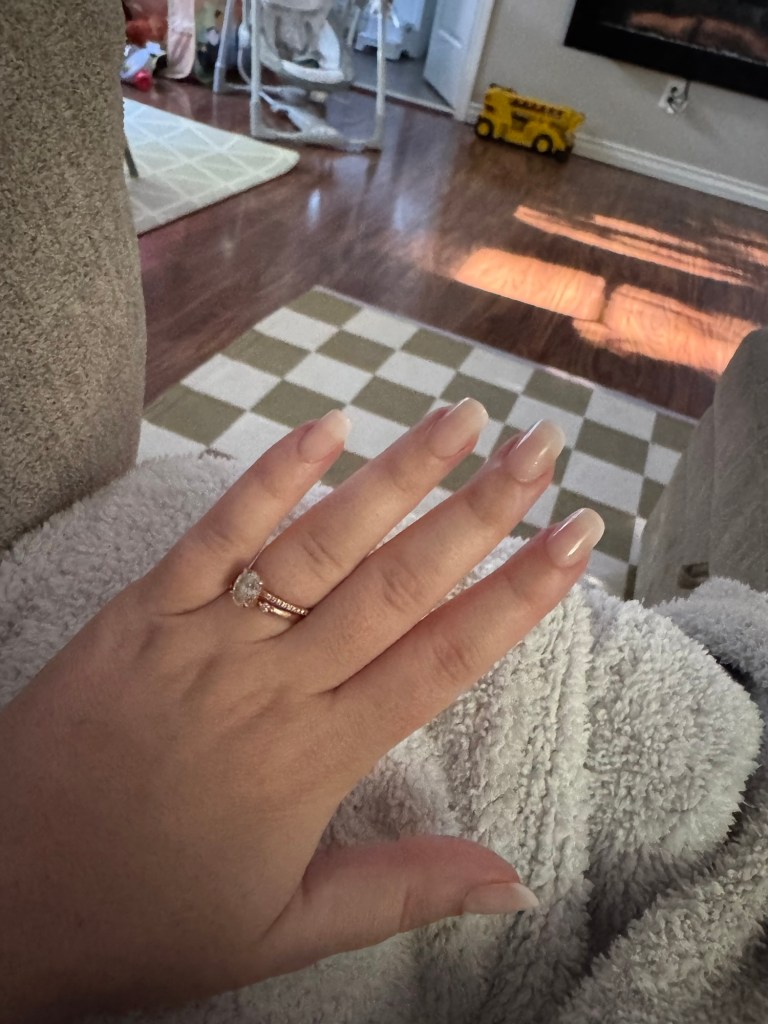 A woman’s hand with nude, glossy press-on nails rests on a soft, light gray blanket. She wears a gold engagement and wedding ring set. In the background, a cozy living room with hardwood floors, a green checkered rug, and children’s toys can be seen, evoking a calm, everyday home setting.